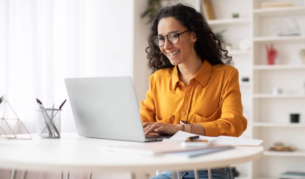 Happy Middle Eastern Businesswoman Using Laptop Typing And Working Online Sitting At Workplace Wearing Eyeglasses. Office Worker Lady Browsing Internet On Computer. Entrepreneurship. Side View