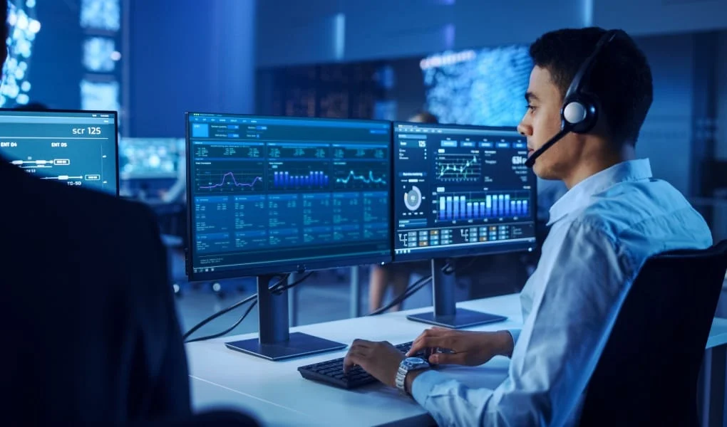 Confident Male Data Scientist Works on Personal Computer Wearing a Headset in Big Infrastructure Control and Monitoring Room. Young Engineer in a Business Call Center Office Room with Colleagues.